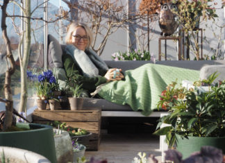 Anne sitter i utesofa med pledd, mellom krukker med planter